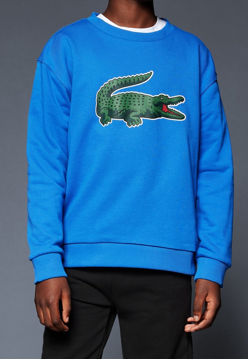 Personne portant un sweat-shirt bleu vif avec un grand crocodile vert dessin animé sur la poitrine, associé à un pantalon noir.