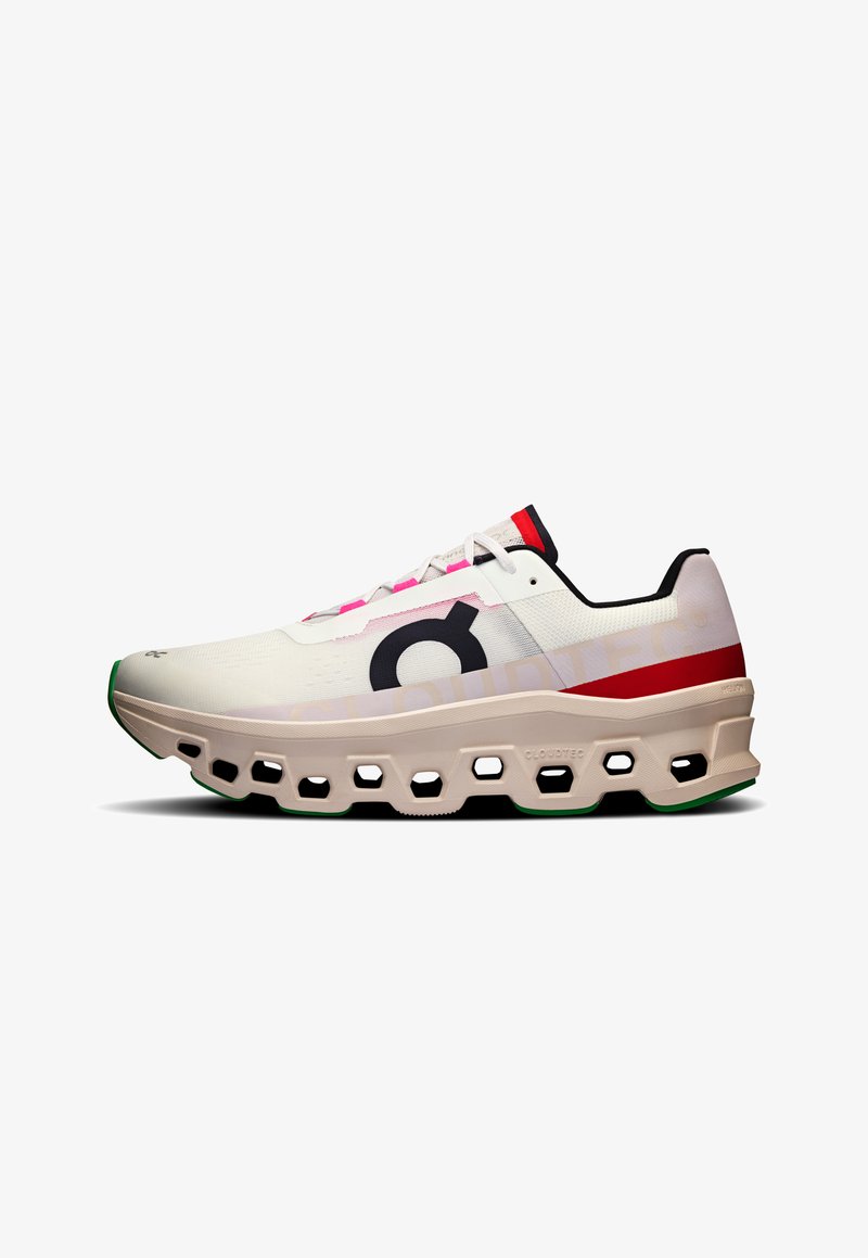 Zapatilla de running con parte superior de malla blanca con acentos rosas, gran logo negro y una suela voluminosa color crema con recortes hexagonales y detalles verdes.