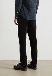 Uomo che indossa pantaloni neri a gamba dritta, scarpe nere con lacci e una camicia a maniche lunghe a quadri blu e verdi, in piedi su un pavimento chiaro.