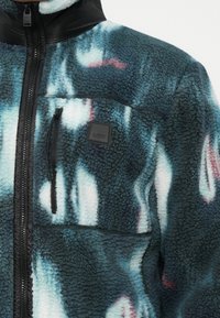 Fleece jacket featuring a blue and white abstract pattern, black zipper, and a small black label. Textured fabric with a zippered pocket.