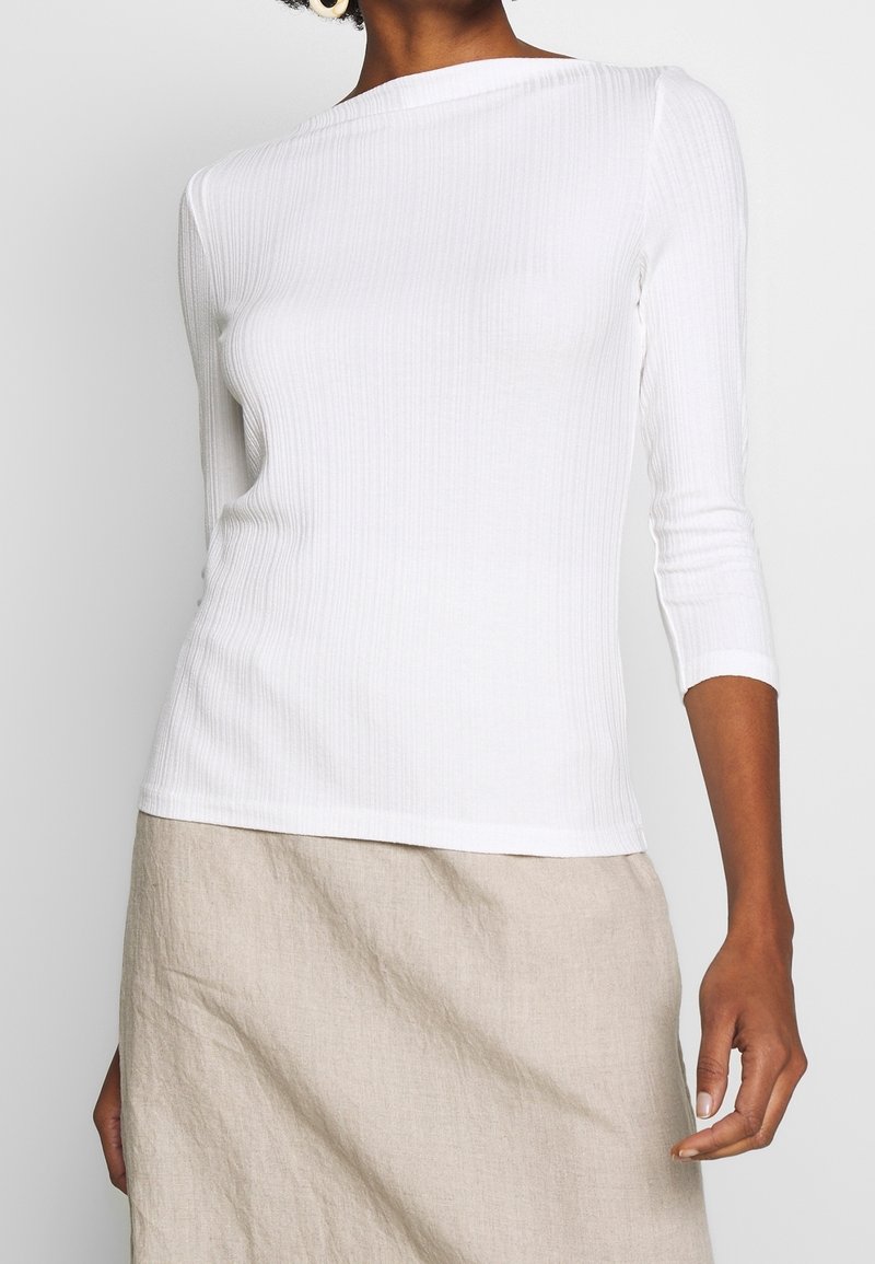 Top blanco de manga larga con canalé y escote barco, diseño ajustado y mangas tres cuartos, combinado con una falda de lino beige.