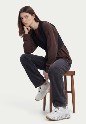 Donna con maglia marrone a maniche lunghe e gilet nero seduta su uno sgabello di legno, indossa jeans scuri e sneaker bianche con suola color gomma.