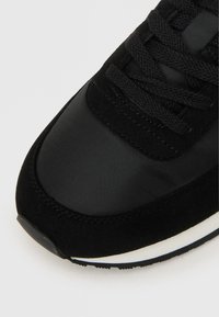 Sapatilha preta com uma combinação de material sintético liso e camurça suave, com atacadores texturizados e um detalhe em sola de borracha branca.