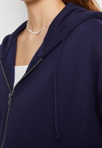 Sudadera con cremallera de color azul marino, con capucha, que presenta una cremallera, cordones y una textura suave. Lleva encima una camiseta blanca. Diseño simple.