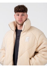 Chaqueta de forro polar beige con un diseño acolchado, cuello alto y cremallera frontal. Presenta un parche con el logo en el pecho. Textura suave, estilo casual.