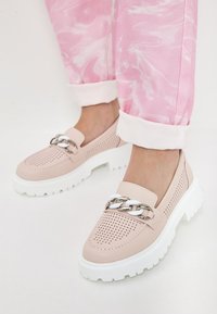Světle růžové perforované slip-on boty s robustní bílou podrážkou a stříbrným řetízkovým akcentem. Nenošeny s ohrnutými růžovými mramorovanými džíny.