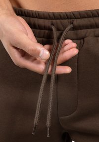 Brązowe bawełniane spodnie dresowe z prążkowanym pasem, ozdobione plecionymi sznurkami i boczną kieszenią. Gładka tekstura i luźny krój.