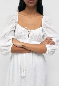 White linen dress featuring a square neckline and puffed sleeves. Ties at the front with tassels at the hem, creating a textured finish.