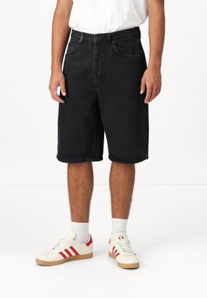 Homme portant un short en denim noir mi-cuisses, des chaussettes blanches montantes, des baskets blanches avec des rayures rouges, et un t-shirt blanc, debout sur un sol blanc.