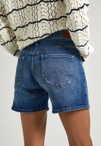 Denim kratke hlače v temno modri barvi, z značilno petimi žepi, zavitim robom in subtilnim zbledelostjo na blagu. Zadnji žepi s podrobnostmi šivanja.