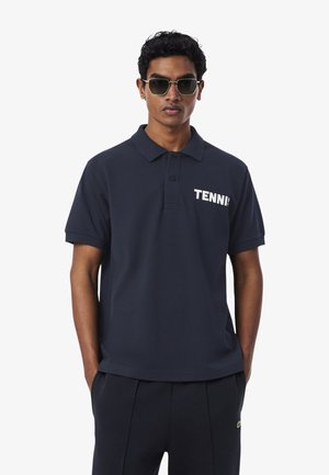 Hombre con polo azul marino oscuro con el texto "TENNIS", pantalones a juego y gafas de sol, de pie con las manos en los bolsillos contra un fondo blanco.