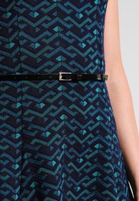 Niebieska i zielona sukienka w wzory z zygzakowymi wzorami, z czarnym paskiem i metalową klamrą, wykonana z teksturowanej tkaniny.