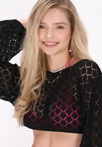 Fekete csipkés crop top gyémántmintával és harangujjakkal, egy rózsaszín felsőre rétegezve. A modellen hosszú szőke haj és többsoros nyakláncok találhatók.