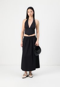 Svart halterneck-top med dyp V-hals, kombinert med en svart maxi-skjørt og beige flate sko. Modellen holder en liten svart håndveske.