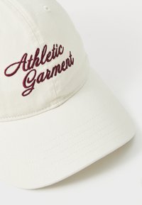 Kremfarget baseballcaps med burgunder brodert tekst som sier "Athletic Garment" på frontpanelet.