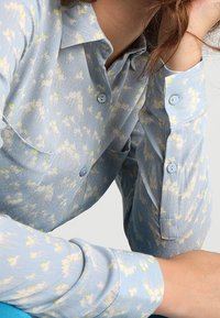 Camicia azzurra chiara con un motivo floreale in bianco e giallo. Presenta maniche lunghe, colletto a punta e due tasche sul petto.