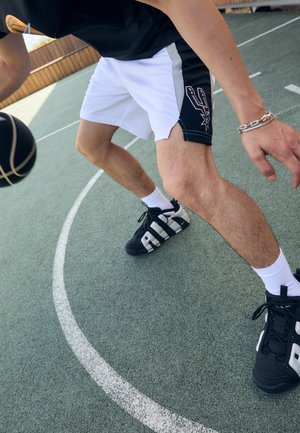 Mężczyzna w czarno-białych szortach do koszykówki kozłuje czarną piłką na zewnętrznym boisku w pobliżu linii za trzy punkty.