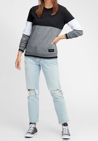 Mujer vistiendo una sudadera de bloques de color negra, blanca y gris, jeans claros rasgados y zapatillas altas negras, de pie con una mano en la cadera.