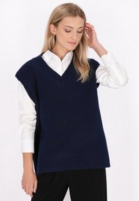 Marineblau gestrickte Weste mit V-Ausschnitt und kurzen Ärmeln, getragen über einem weißen Hemd mit Knöpfen, mit entspannter Passform und weicher Textur.