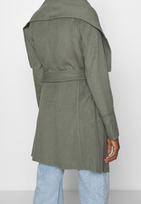 Trench coat verde oliva con ampi revers, vita cintrata e taglio sartoriale. Realizzato in un tessuto liscio e morbido con lunghezza fino a metà coscia.