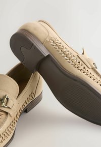 Mocassins en daim beige avec détails tressés sur le côté, boucle en métal et semelles en caoutchouc brun foncé texturées. Une chaussure debout, l'autre montrant la semelle.
