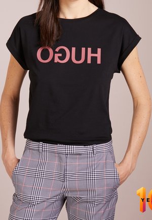 Kobieta w czarnej krótko skrojonej koszulce z odwróconym na czerwono napisem „HUGO” i w kratę spodniach z wysokim stanem, ręce w kieszeniach.