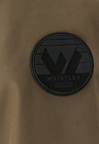 Khaki tyg med en cirkulär svart gummiapplikation med upphöjd text och en stiliserad "W"-logotyp, placerad mot en texturerad bakgrund.