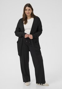 Cardigan en tricot noir à devant ouvert, poignets côtelés et deux poches avant, associé à un haut blanc et un pantalon noir. Texture lisse, coupe décontractée.