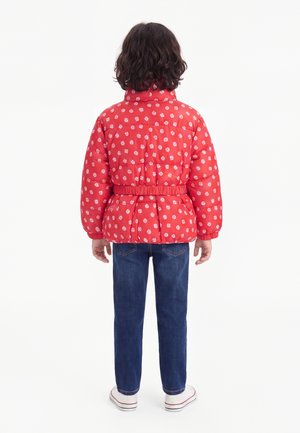Enfant aux cheveux sombres mi-longs portant une doudoune rouge à fleurs, un jean bleu et des baskets blanches, debout de dos.
