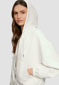 Bijela zip-up dukserica s velikom kapuljačom, prednjim dzepovima i elastičnim manžetama. Mekan materijal s glatkom teksturom i minimalističkim dizajnom.