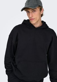 Schwarzer Hoodie mit Kängurutasche aus weichem Material. Verfügt über eine großzügige Kapuze und eine lockere Passform. Getragen mit einer grauen Mütze.