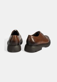 Bruine leren schoenen met een gladde textuur, ronde vorm en een dikke zwarte zool. Bevatten minimale accenten en hebben een vetersluiting.