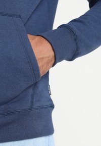 Sudadera de color azul oscuro con un bolsillo tipo canguro. Fabricada en una suave tela con puños de canalé y una etiqueta con el nombre de la marca visible.