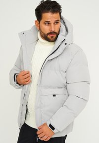 Hellgraue Puffjacke mit einer Kapuze, durchgehendem Reißverschluss und zwei Seitentaschen. Hergestellt aus glattem, wasserabweisendem Material mit verstellbaren Bündchen.