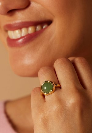 Close-up of a smiling person wearing a gold ring with a round green stone on their finger near their lips.