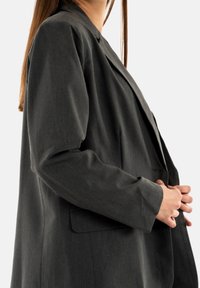 Blazer gris oscuro de tejido texturizado, con cierre de un único botón, dos bolsillos y un corte entallado con hombros estructurados.