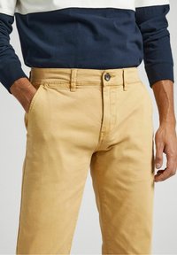 Pantalon chino kaki à texture lisse, coupe droite, poches avant et braguette à boutons, associé à une chemise manches longues bleu marine et blanche.