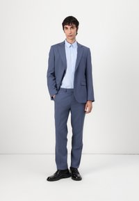 Hellblauer Anzug mit passender Hose und Hemd mit Knöpfen. Schlankes Design, strukturierter Stoff und schwarze Dressschuhe. Schlichte, moderne Ästhetik.