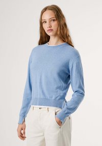 Svetlo modra cashmere pulover s okroglim ovratnikom, rebrastim robom in manšetami. Vsebuje subtilno vezeno logo na levi strani.