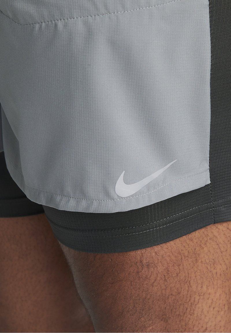 Gros plan sur une jambe à la peau foncée portant un short Nike gris clair avec une couche de compression gris plus foncé en dessous, montrant le logo Nike.