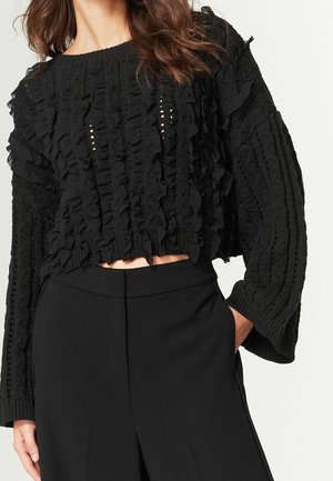 Femme portant un pull court noir avec des volants et des manches larges en maille texturée, associé à un pantalon noir taille haute, main dans la poche.