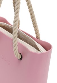 Rosa plastikkasse med tvinnade rephandtag, med ett ljusfärgat foder och en dragkedjeficka. O bag-logotyp på utsidan.