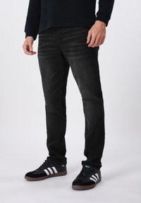 Μαύρο τζιν με λεπτή εφαρμογή και διακριτή κάθετη υφή. Συνδυασμένο με μαύρα αθλητικά παπούτσια με λευκές ρίγες.