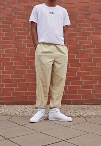 Camiseta blanca de algodón con un pequeño logo negro, pantalones sueltos beige y zapatillas altas blancas, todo colocado frente a un fondo de ladrillo.