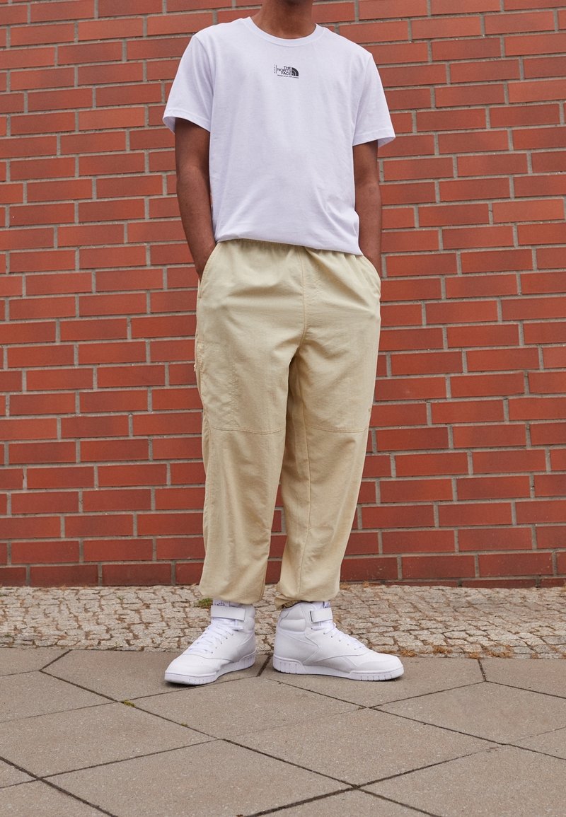 Camiseta blanca de algodón con un pequeño logo negro, pantalones sueltos beige y zapatillas altas blancas, todo colocado frente a un fondo de ladrillo.