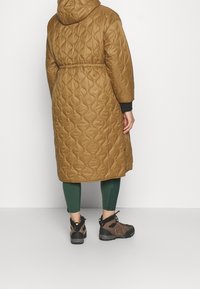Abrigo largo acolchado marrón con capucha, cintura ceñida y bolsillos laterales, combinado con leggings verdes y botas de senderismo marrones.