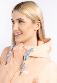 Impermeabil de culoare piersică cu guler înalt, șireturi albastre și albe în dungi, mărgele din lemn și un logo cu umbrelă albă pe piept.