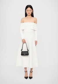 Witte off-the-shoulder jurk met een gestructureerde lijfje en lange mouwen, gecombineerd met een zwarte leren handtas en puntige hakken.