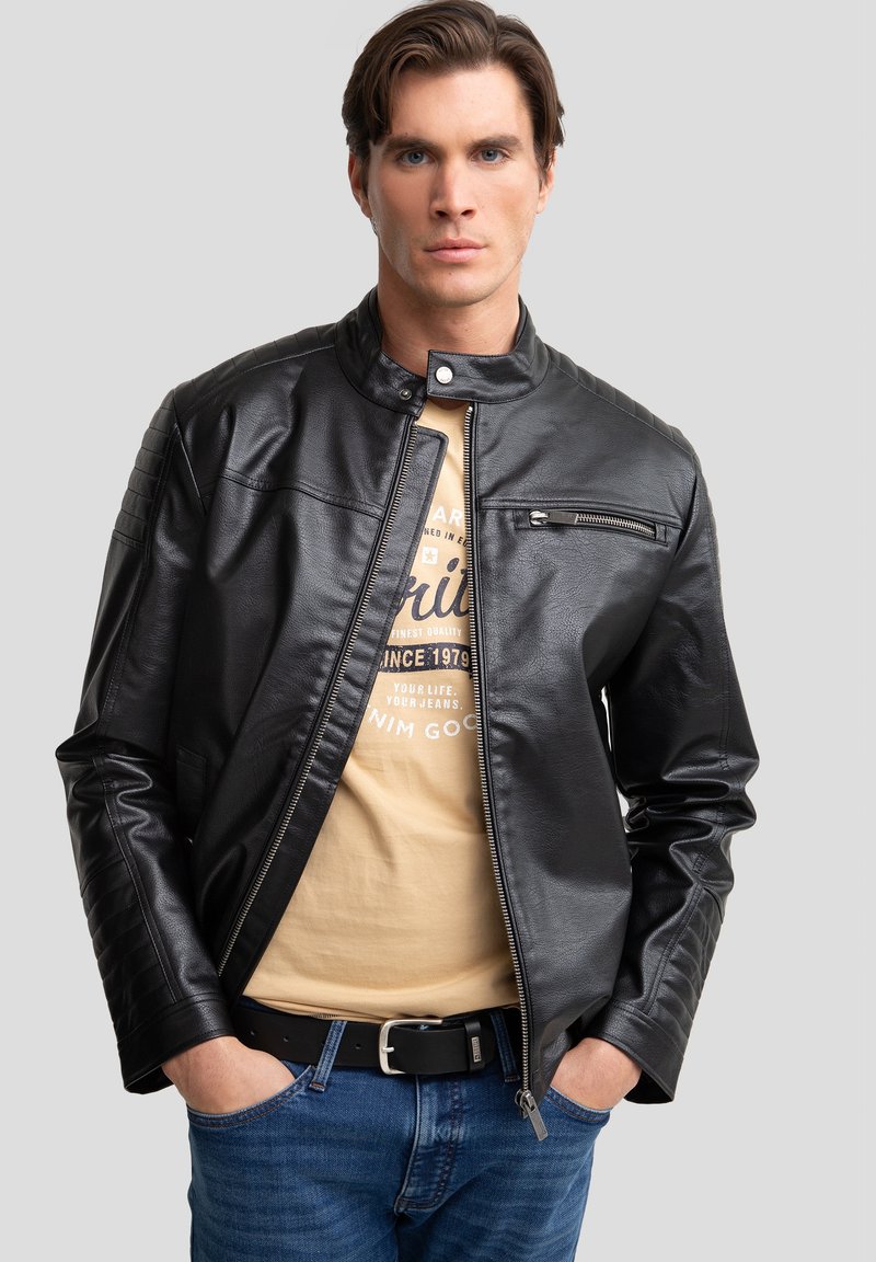Zwarte leren jas met een staande kraag, ritssluiting aan de voorkant en geribbelde schouderdetails, gedragen over een beige shirt en blauwe spijkerbroek.