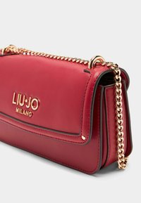 Červená kožená kabelka s zlatým řetízkovým popruhem. Má klopnu na přední straně, embosované logo a kontrastní prošívání podél okrajů. Kompaktní design.